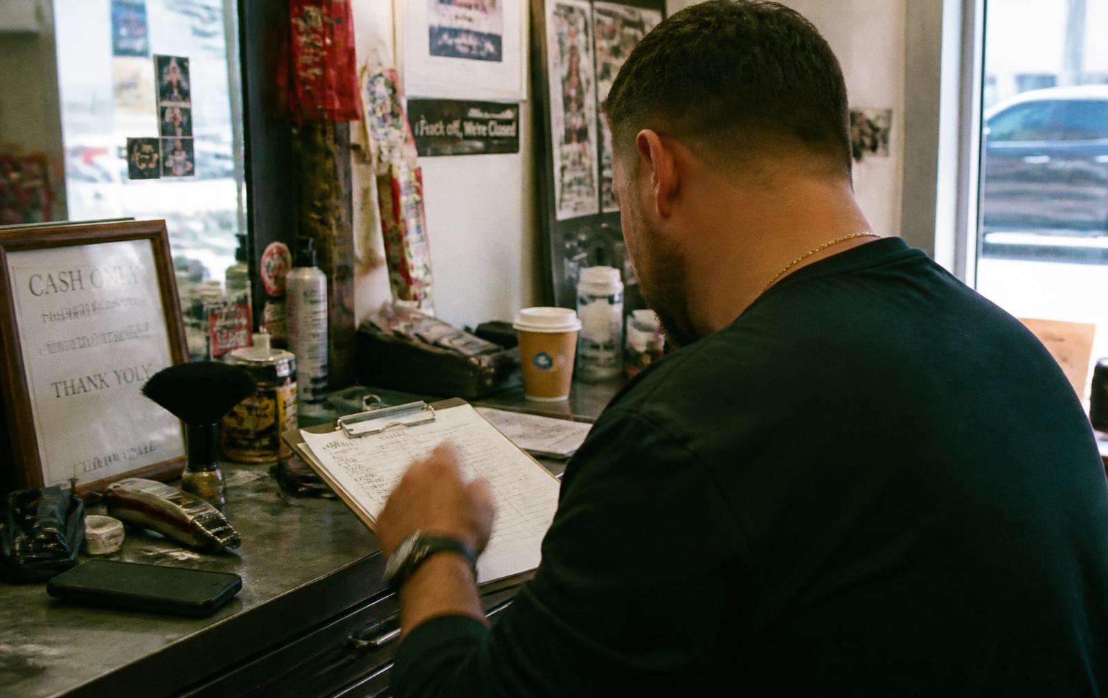 A barbershop owner at the counter, looking over a handwritten client list on a clipboard