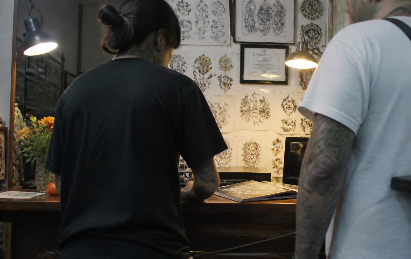 A tattoo artist at the front desk with a returning client, flash sheets on the wall behind